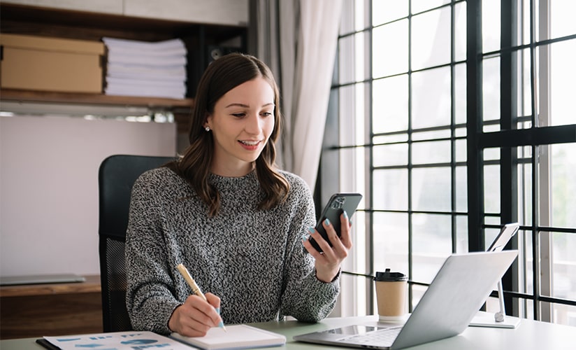 Femme écrivant dans un agenda tout en tenant un téléphone intelligent et en parcourant son ordinateur portable